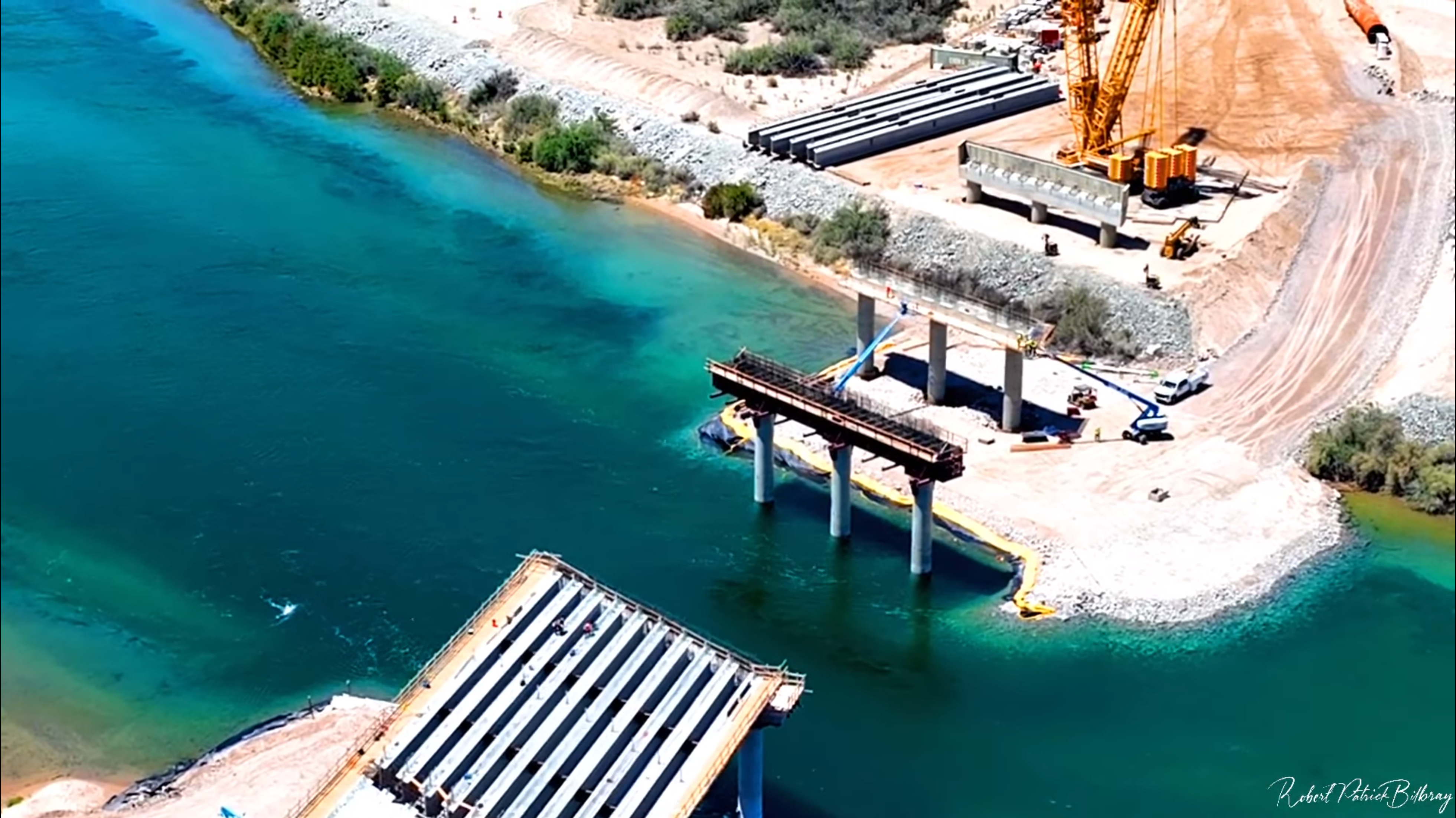 Aerial view of the Laughlin-Bullhead City Bridge Project under construction. In the foreground, cranes and some pilons can be seen. The Colorado River flows beneath the bridge, with the desert landscape in the background.
